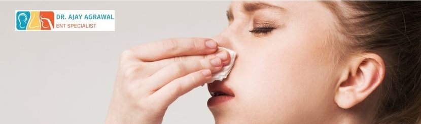 Here is the analysis of the image: ALT TEXT: A woman holds a tissue to her nose, with her eyes closed, appearing to be dealing with a nosebleed. The logo and name of Dr. Ajay Agrawal, ENT Specialist, are included.
